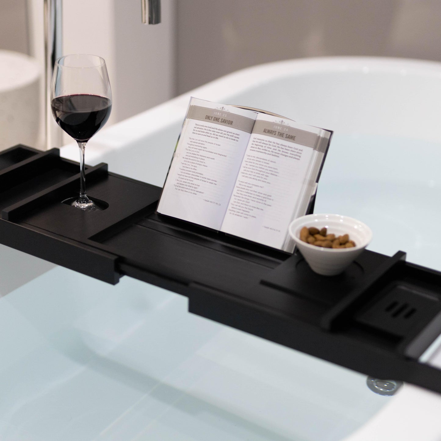 Black bamboo bath caddy with wine glass, open book, and snack bowl on tray