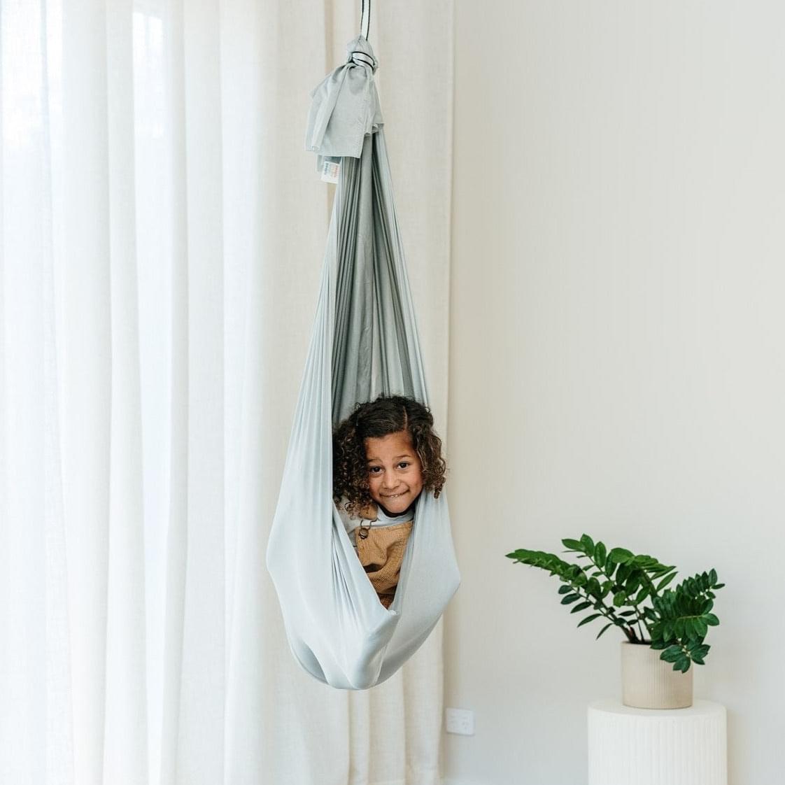 Light grey therapy swing cocoon hanging from above, child seated inside in draped fabric