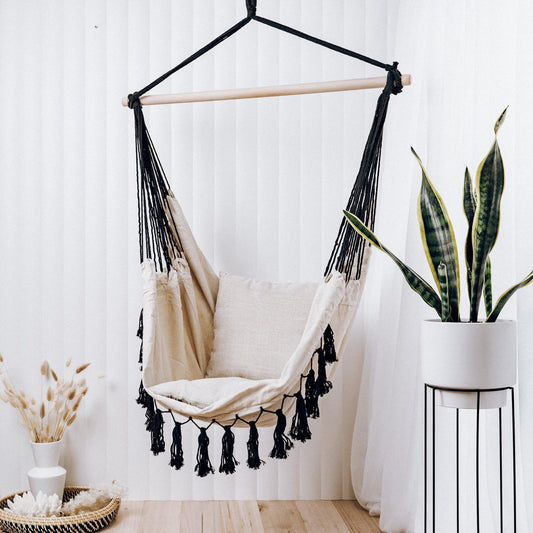 Cream hammock chair with black tassel fringe, suspended frame, and seat cushion.