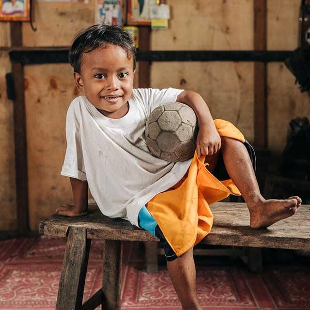 Child in white shirt holding a soccer ball with orange cloth draped over legs.