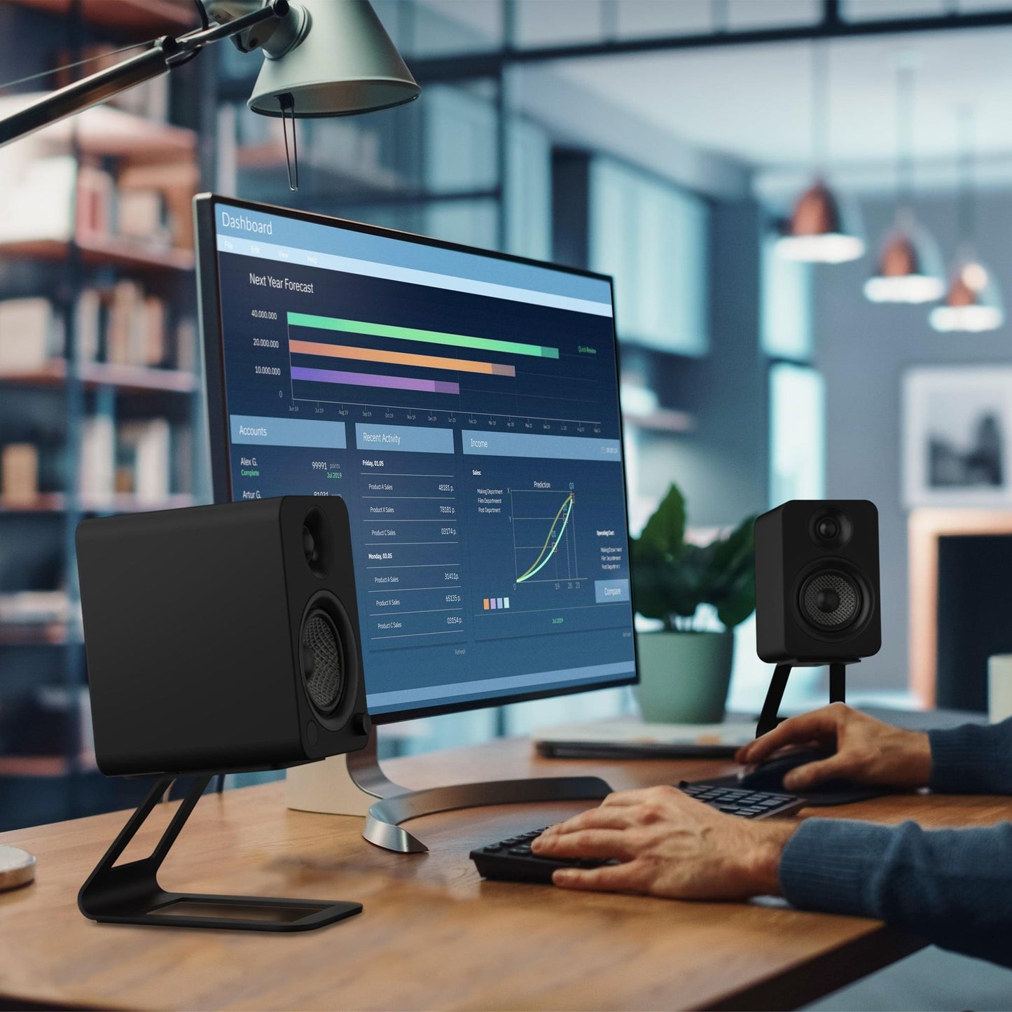 Speakers on black elevated desktop stands beside a monitor in a workspace