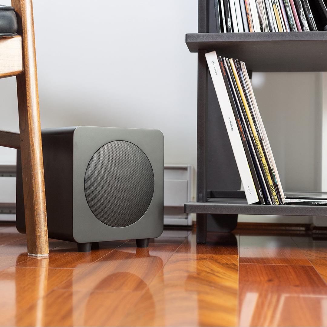 Matte black powered subwoofer side view with circular grille, on the floor