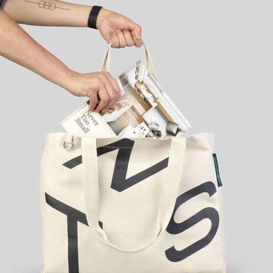 Cream canvas tote bag open with hands inside, black monogram front panel