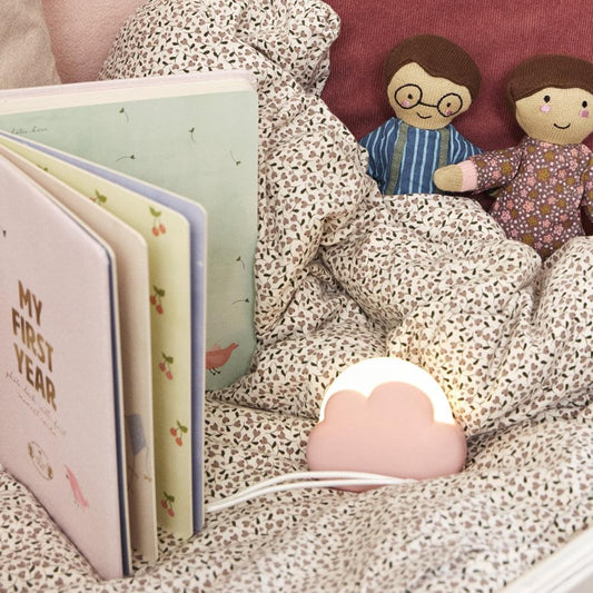 Glowing dusty rose cloud-shaped night light on a bed near books and dolls