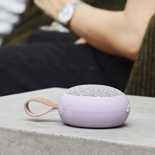 Compact lilac fabric Bluetooth speaker resting on surface, showing side buttons and strap.