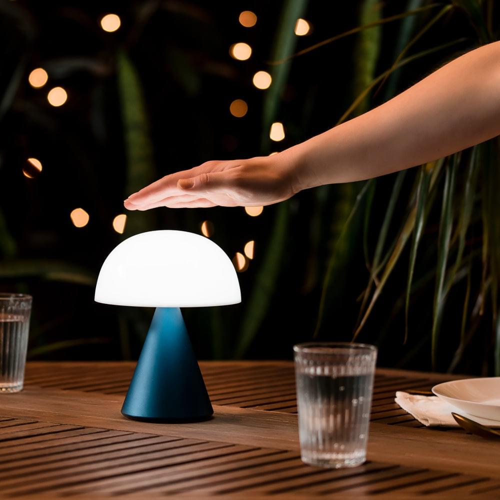 Portable LED lamp with white dome shade and blue base, hand reaching nearby
