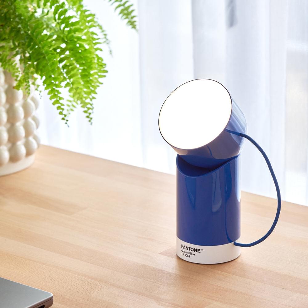 Blue portable LED lamp with glowing round head at angle, white base, PANTONE logo, cord