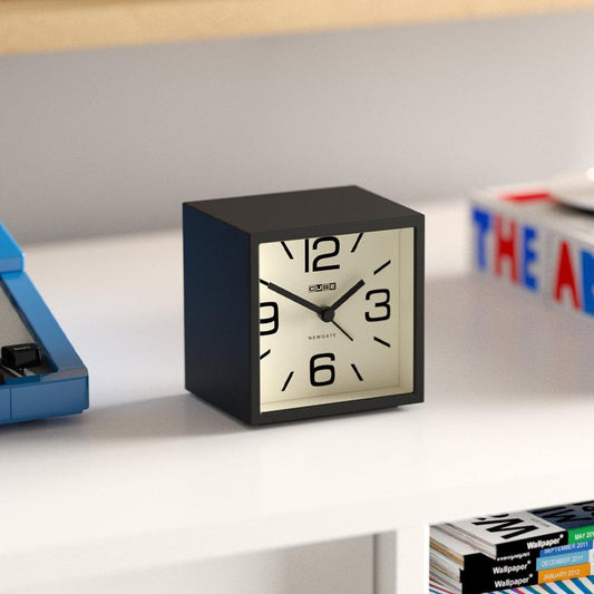 Black cube mantel clock with cream dial and bold numerals on a shelf.