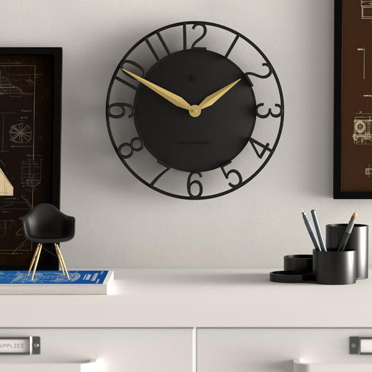 Black wall clock on wall with gold hands and decorative number ring, centered face.