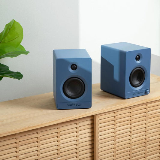 Two blue bookshelf speakers with black woofers and tweeters on wooden shelf