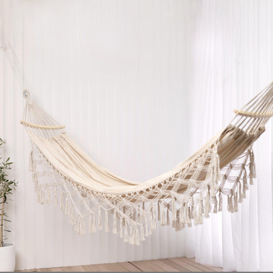 Cream hammock with woven net and long tassel fringe, between wooden spreaders