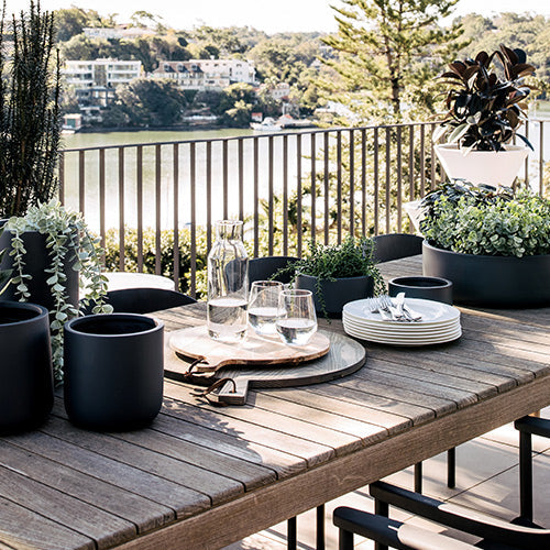 Three dark round planters with green plants on an outdoor wooden table