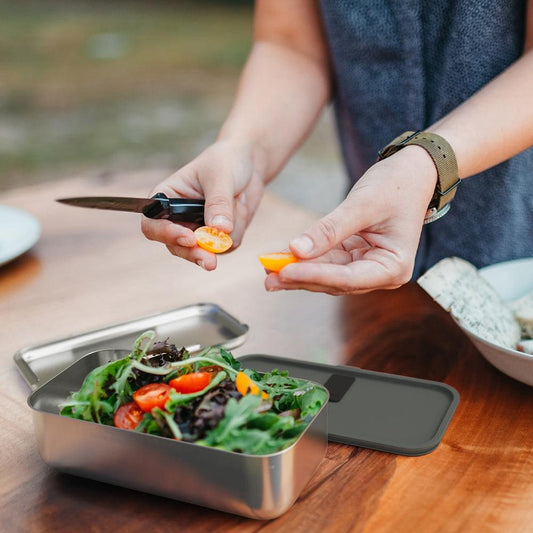 Open stainless bento box with orange strap; hands placing food inside