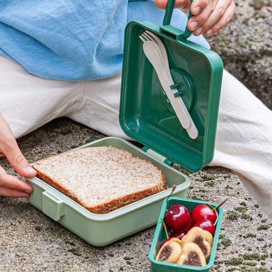 Green lunch box open with sandwich in base and snack compartment; hands holding lid